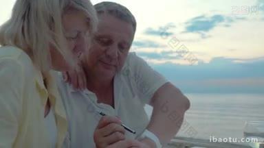 老年夫妇使用<strong>智能手表</strong>坐在海滩咖啡馆在晚上男子触摸屏幕与笔和显示一些东西